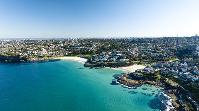 Aerial view of a coastal town with turquoise ocean, sandy beaches, and houses on hills under a clear blue sky.