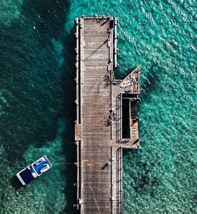 Aerial overhead shot of a long wooden jetty extending into clear turquoise ocean water. A white and blue boat is tied to the jetty. Several people are visible on the jetty, some sunbathing on a platfo