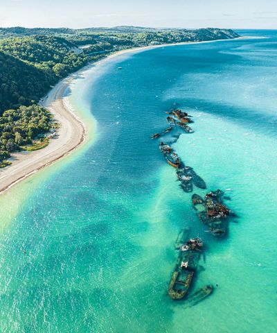 Aerial view of Tangalooma Wrecks, a line of partially submerged shipwrecks in clear turquoise ocean water next to a white sandy beach and lush green coastline under a bright sky.