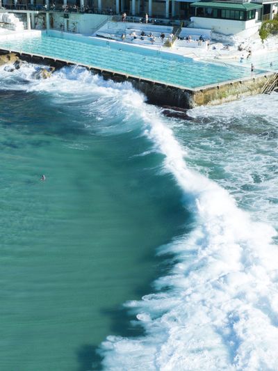 Aerial view of the iconic Bondi Icebergs Club ocean pool in Sydney, Australia. People swim and sunbathe. Powerful turquoise waves crash against the pool's edge, creating a dynamic contrast with the ca