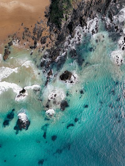 Aerial view of a beach with golden sand, dark rocks, and clear turquoise ocean water with white waves breaking on the shore.