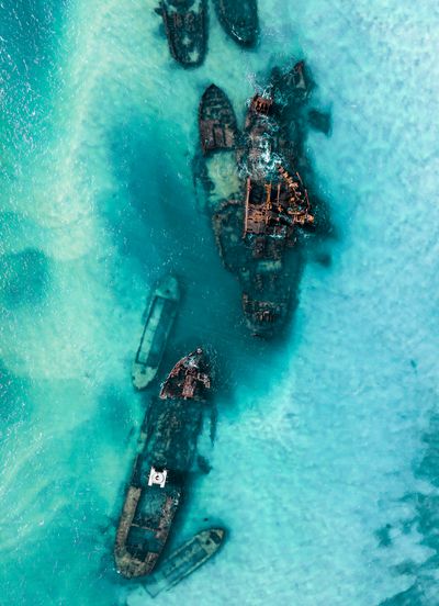 Aerial top-down view of multiple rusty shipwrecks partially submerged in clear, shallow turquoise ocean water. Waves gently lap around the decaying vessels.