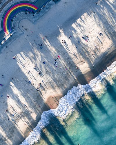 Aerial view of Bondi Beach showing a rainbow-colored walkway, sandy beach with many people and long shadows, and turquoise ocean with white waves crashing on the shore.