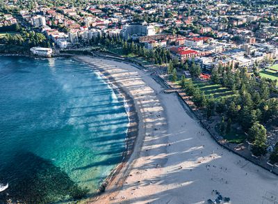 Aerial view of a curved sandy beach with turquoise water and breaking waves. People are scattered on the beach and swimming. A coastal town with buildings and trees lines the background, casting long 