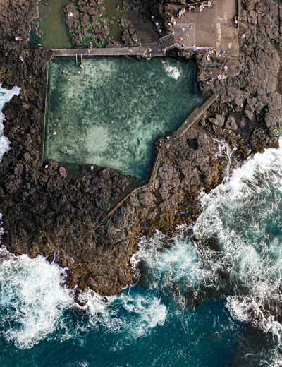 Aerial view of a natural ocean swimming pool carved into dark volcanic rock, with several people swimming and sunbathing. Waves crash against the rugged coastline in the foreground.