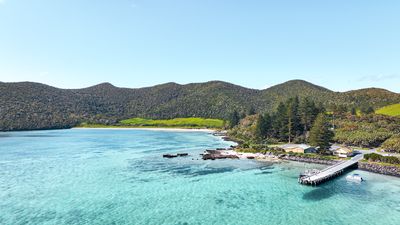 Aerial view of a pristine bay on Lord Howe Island with clear turquoise water, a sandy beach, dense green hills, and a wooden jetty with a small boat.
