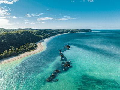 Aerial view of a line of shipwrecks partially submerged in clear turquoise ocean water, alongside a white sandy beach and green forested hills under a blue sky with clouds.